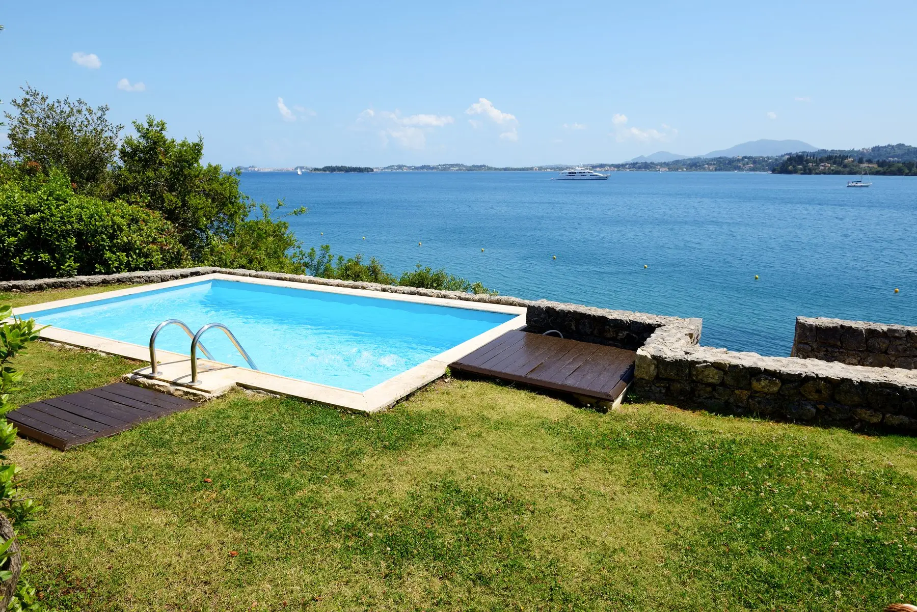 Une piscine bordée de pelouse, au bord d’un muret de pierre qui domine l’eau