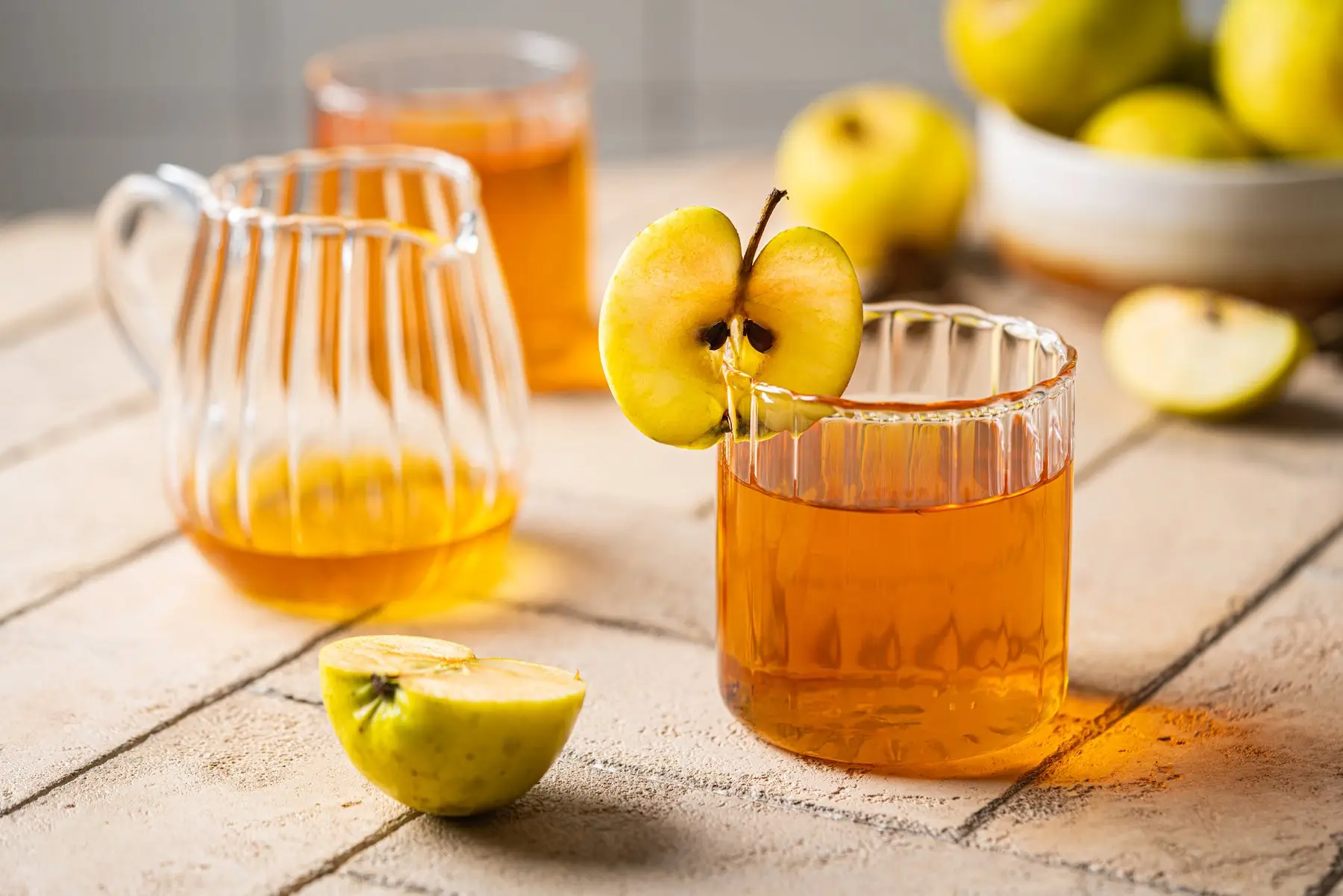 Pichet et verre de cidre doré sur une table claire, avec des pommes jaunes coupées