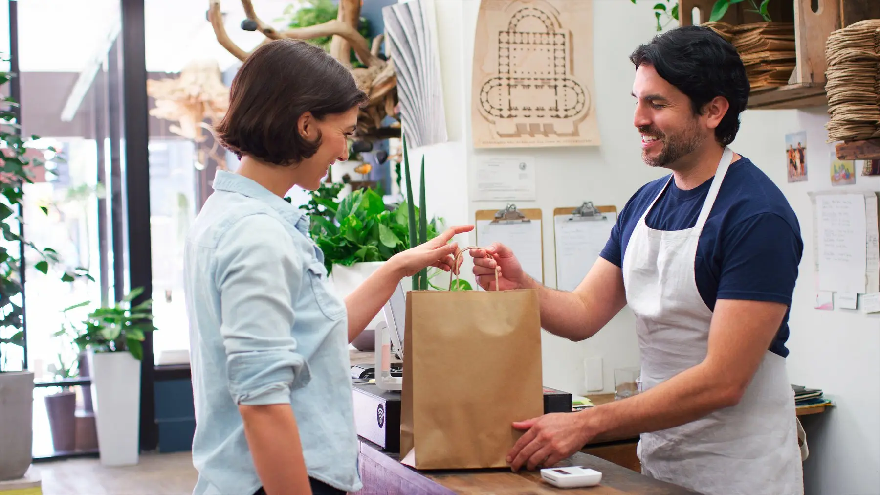 Un commerçant en tablier tend un sac en papier kraft à une cliente, au comptoir de sa boutique
