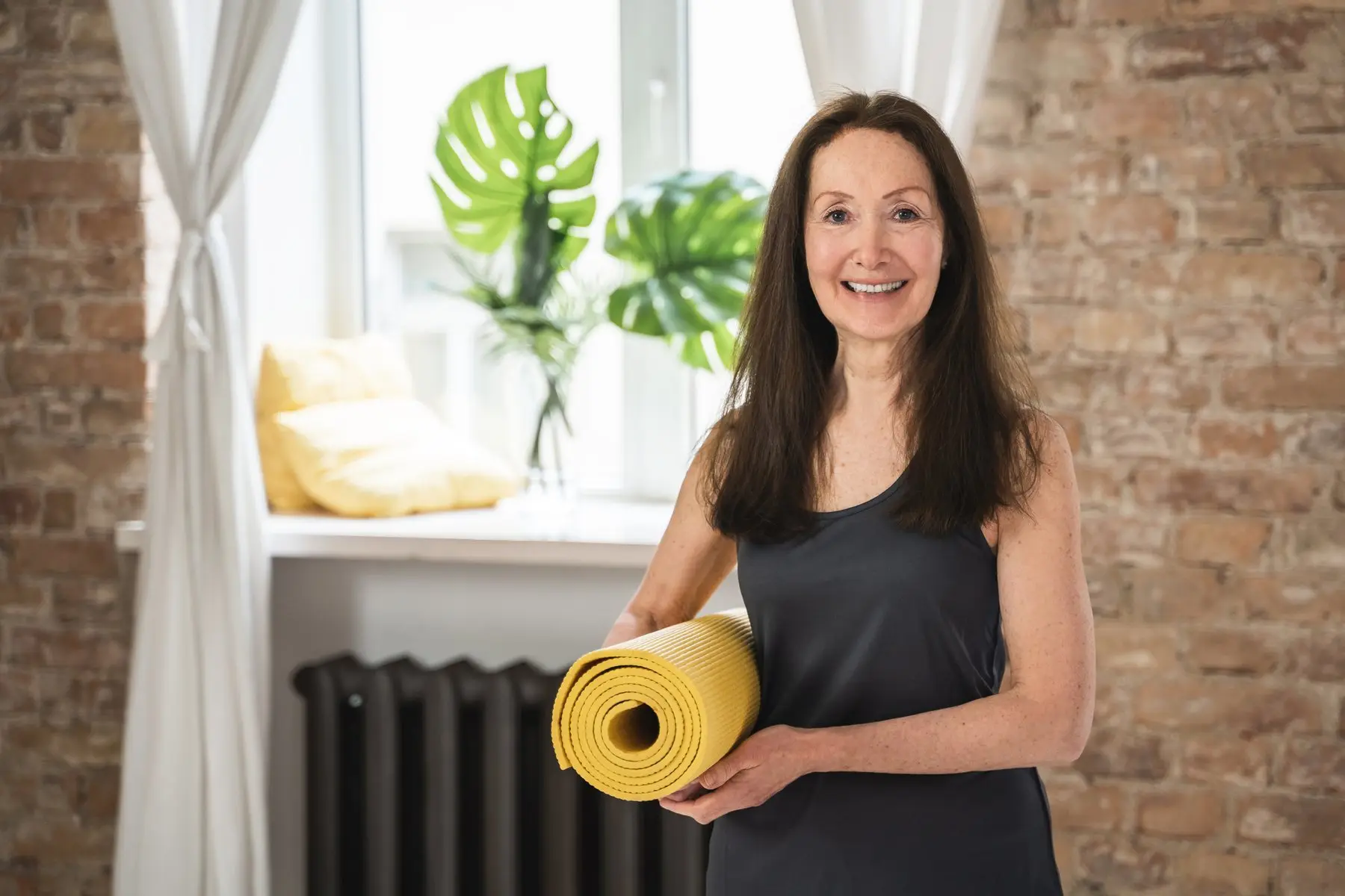 Une femme souriante tient un tapis de yoga jaune roulé, dans une pièce lumineuse aux murs de briques