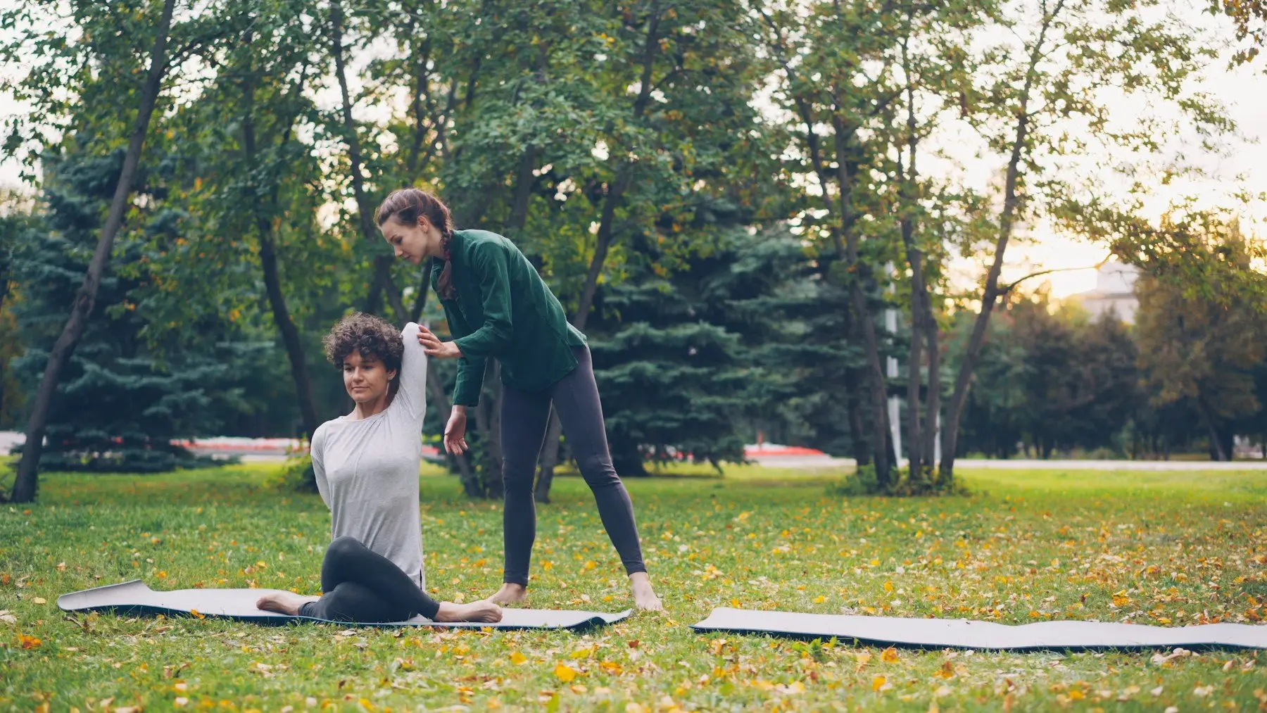 Une professeure ajuste la posture d’une élève assise sur un tapis, dans un parc arboré semé de feuilles mortes