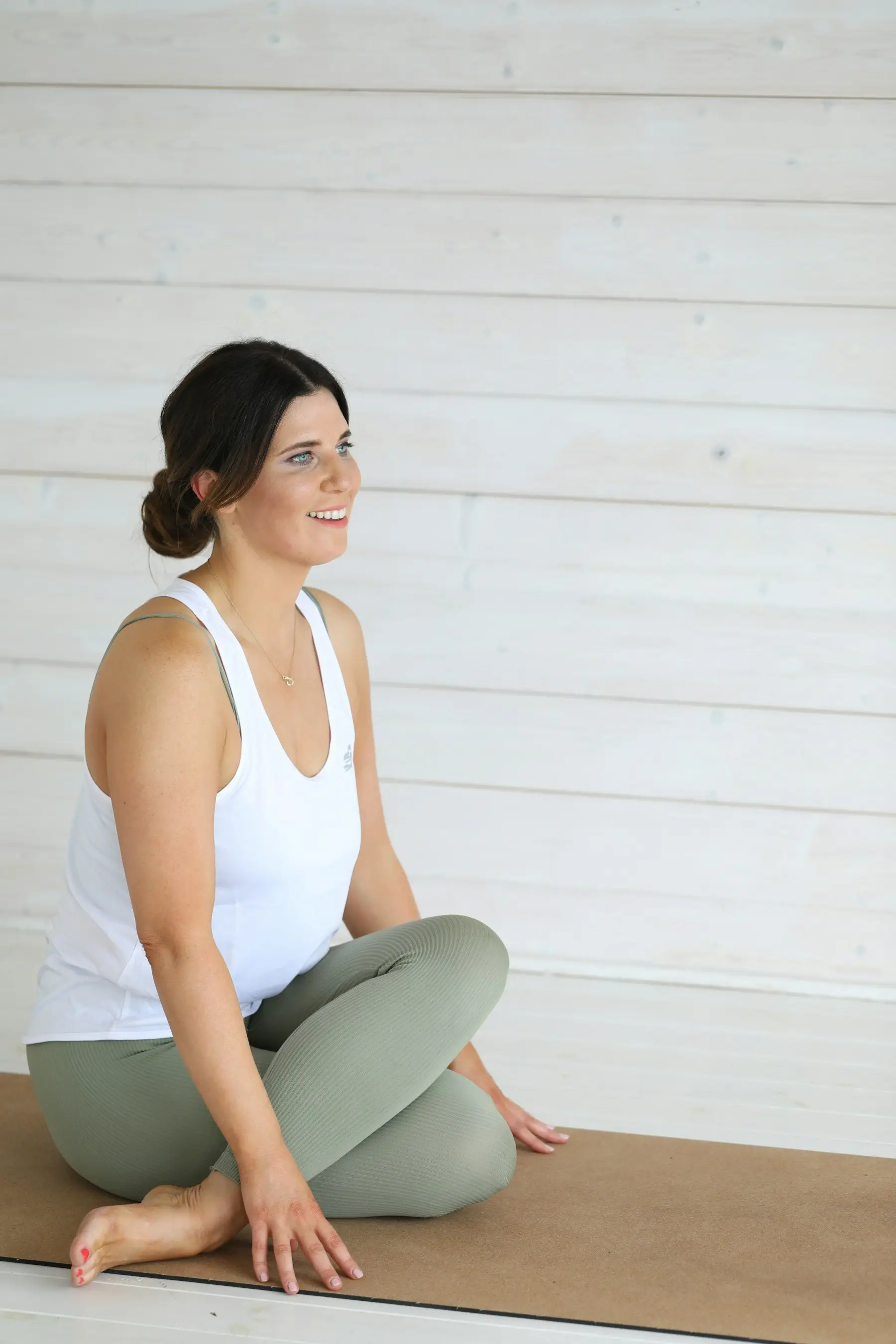 Une professeure de yoga souriante, assise jambes croisées sur un tapis en liège, devant un mur de lambris blanc