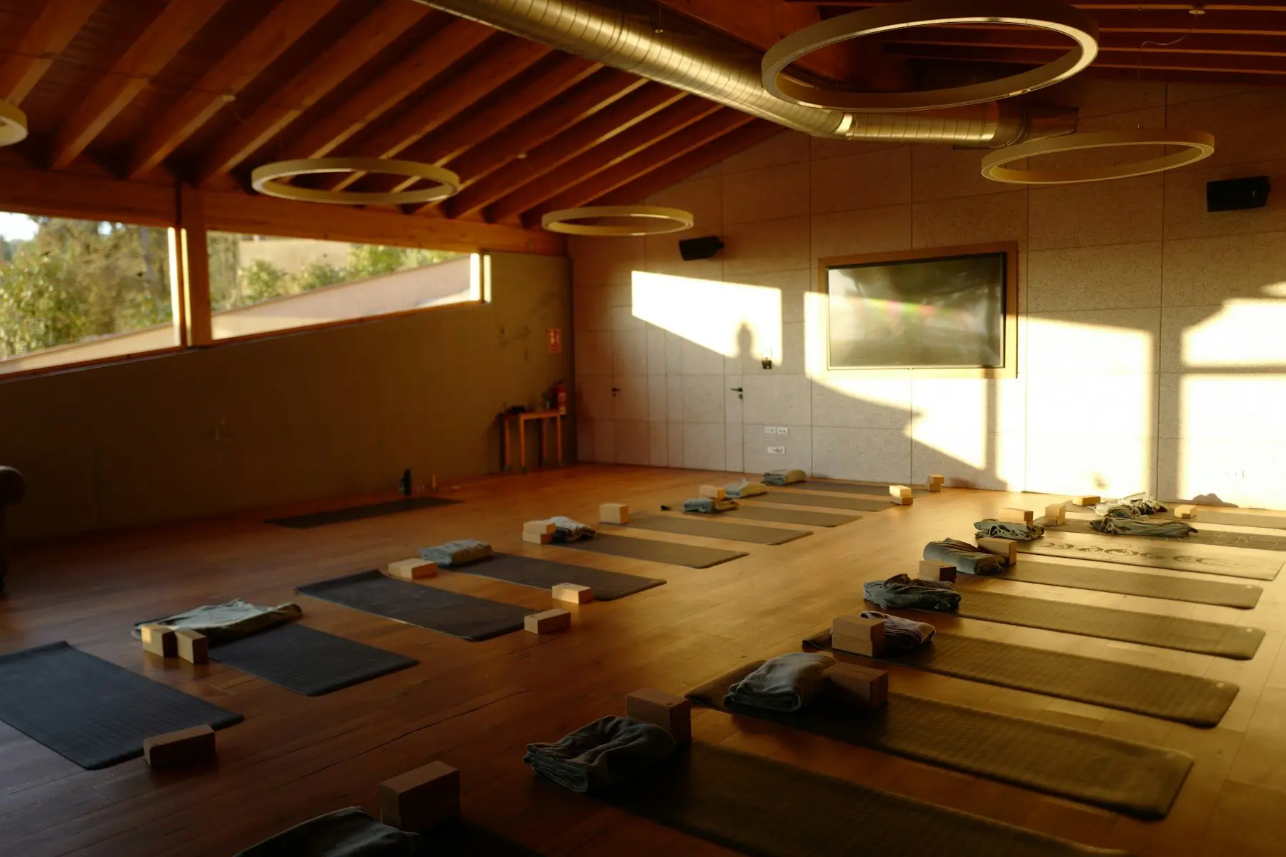 Salle de yoga sous une charpente en bois, tapis alignés avec briques et couvertures pliées, lumière de fin de journée sur le mur