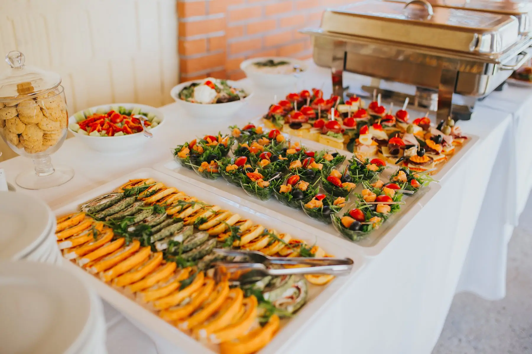 Buffet sur nappe blanche : plateaux de roulés, de canapés et de brochettes de tomates cerises, un chauffe-plat en arrière-plan