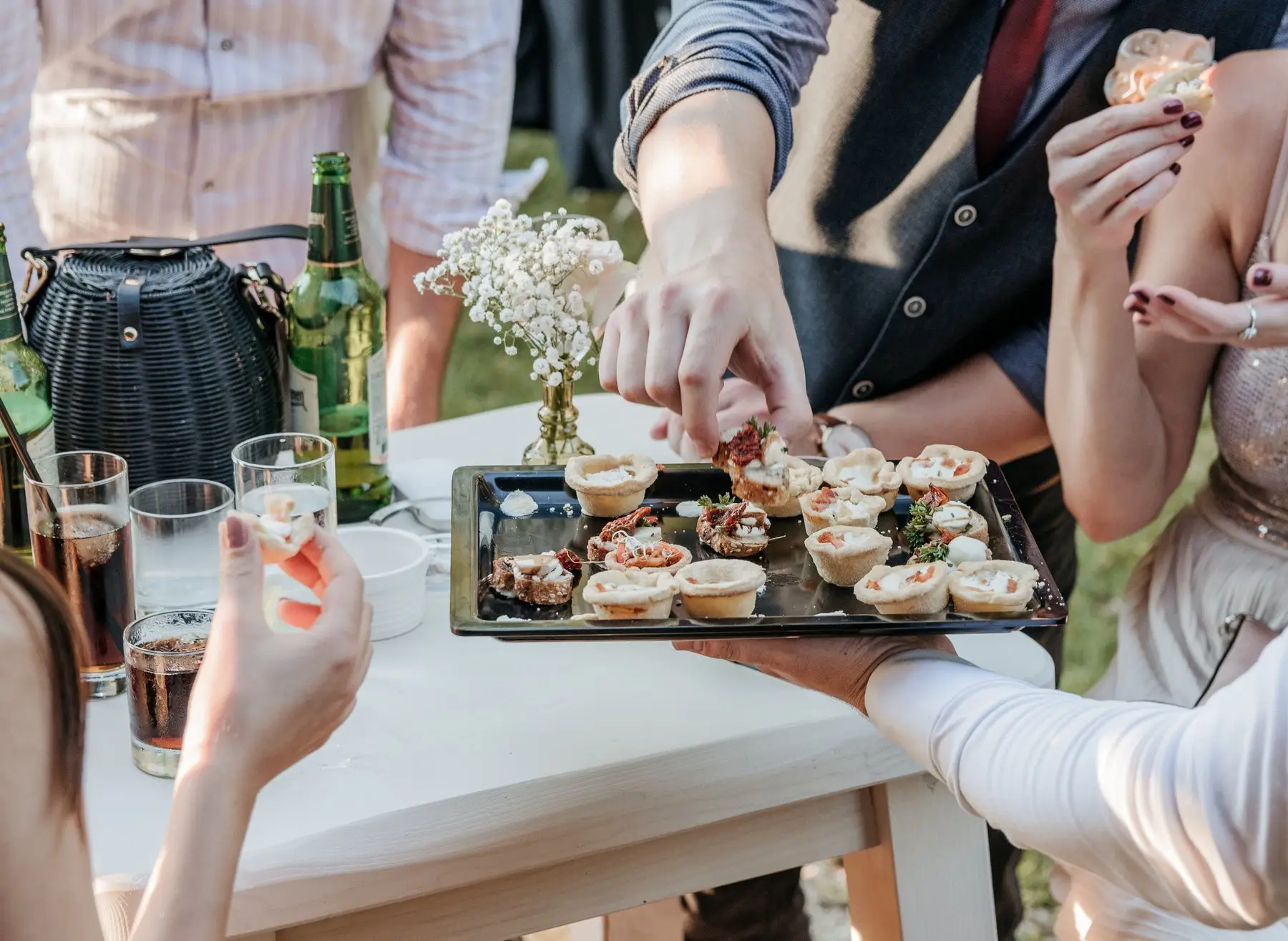 Des invités se servent en mini-tartelettes sur un plateau tendu au-dessus d’une table blanche dressée en plein air