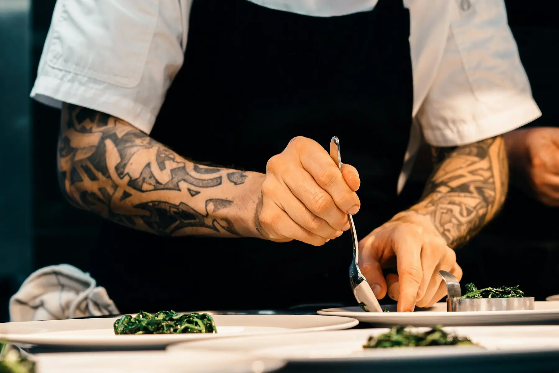 Un cuisinier aux avant-bras tatoués dresse des assiettes à la cuillère, en tablier noir