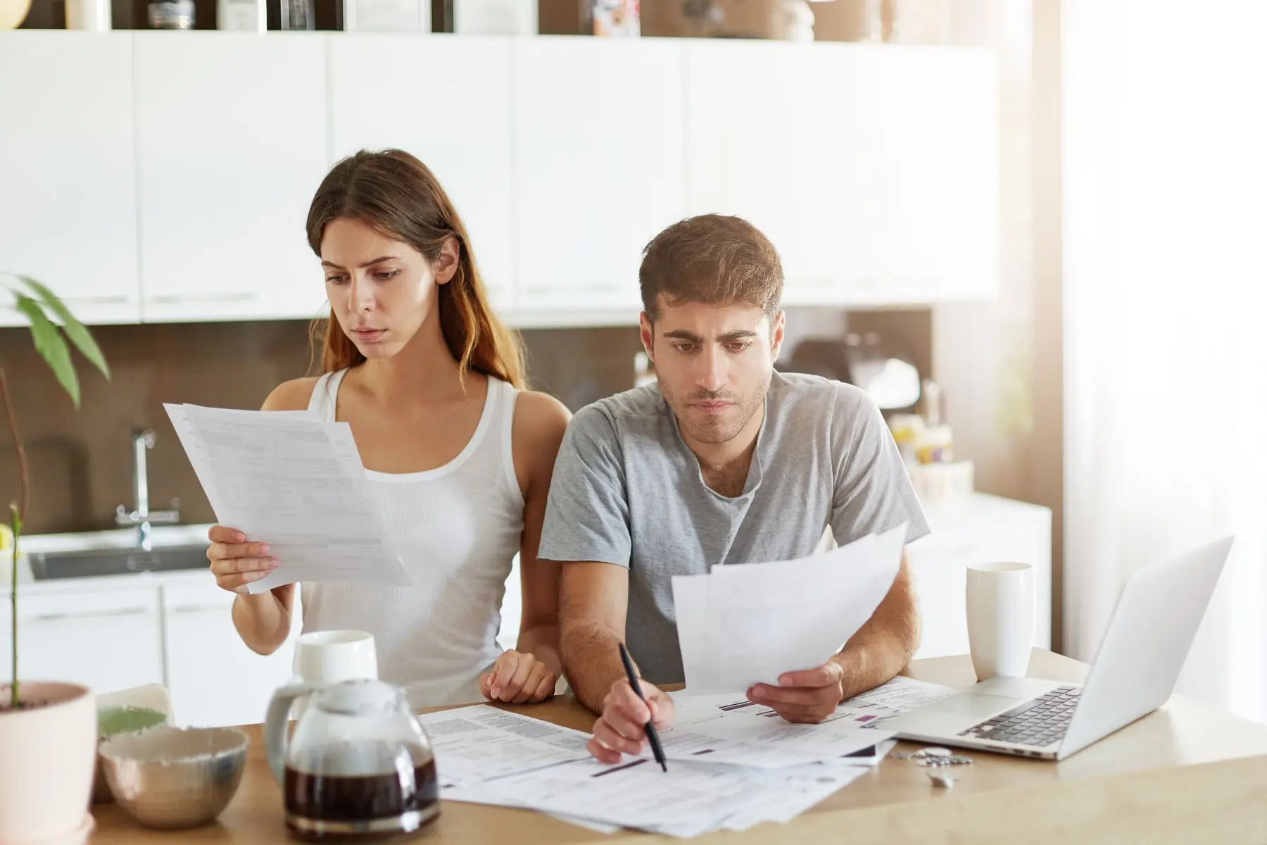 Un couple étudie attentivement des documents à la table de sa cuisine, à côté d’un ordinateur portable