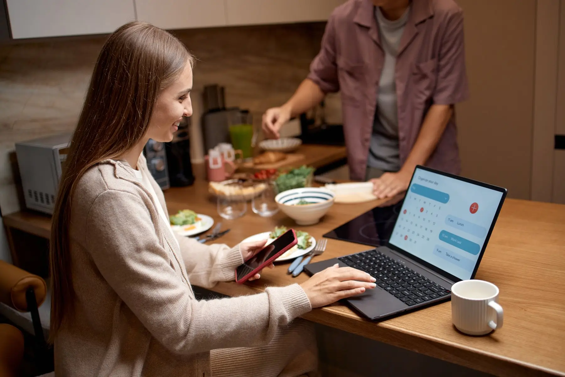 Une femme souriante utilise son ordinateur portable et son smartphone à la table de sa cuisine
