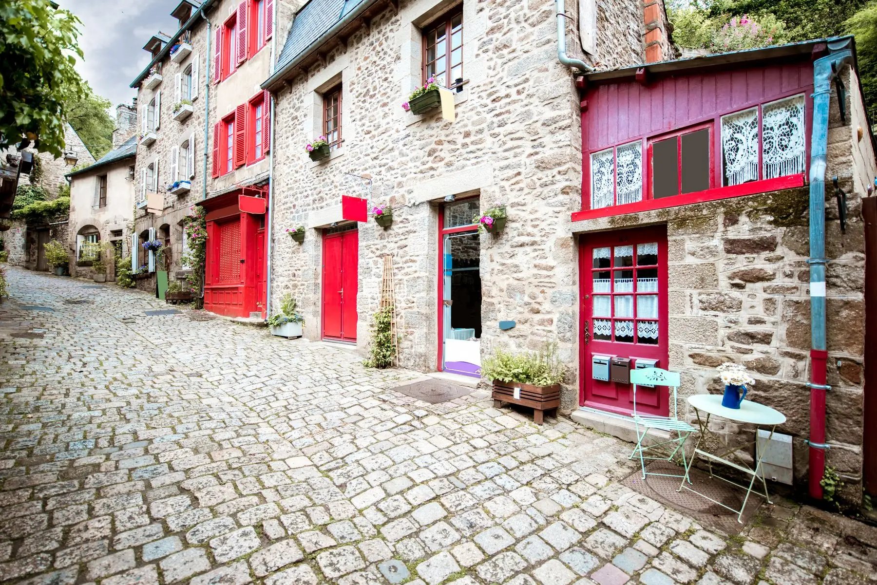 Ruelle pavée en pente bordée de maisons en pierre aux portes et volets rouges, avec une petite table de bistrot devant une entrée
