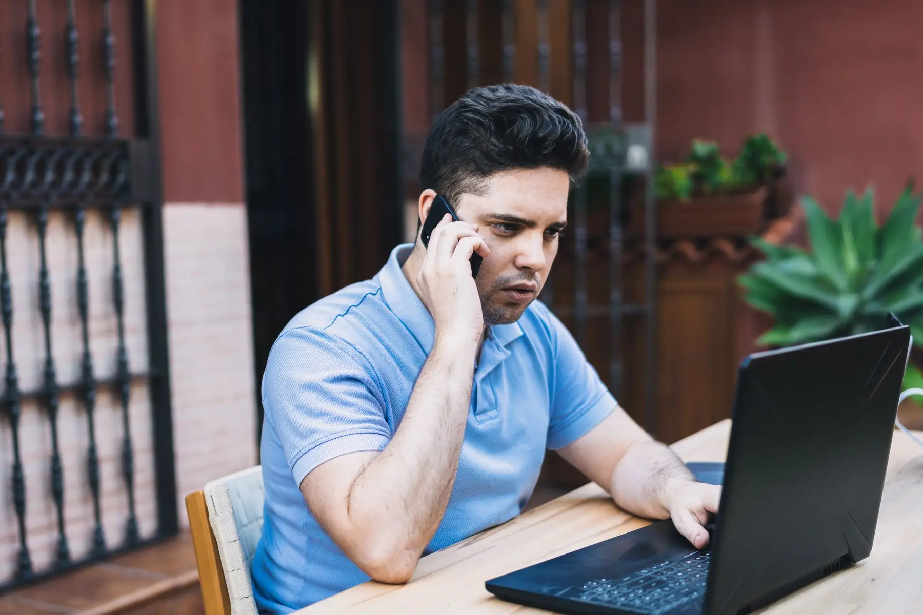 Un homme au téléphone, la main sur le clavier de son ordinateur portable, assis à une table en extérieur