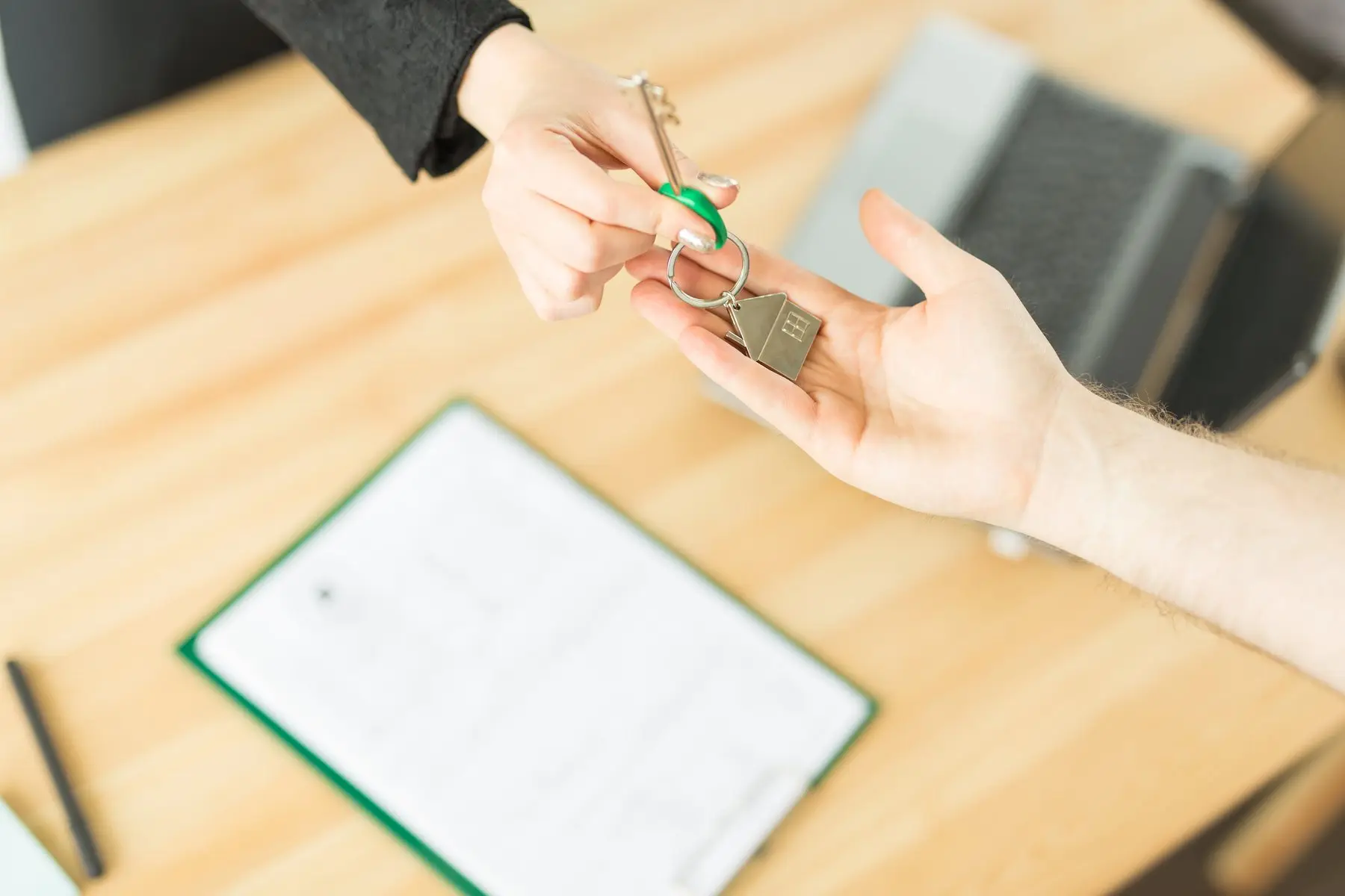 Une main tend un trousseau de clés à porte-clés en forme de maison à une autre main, au-dessus d’un bureau en bois