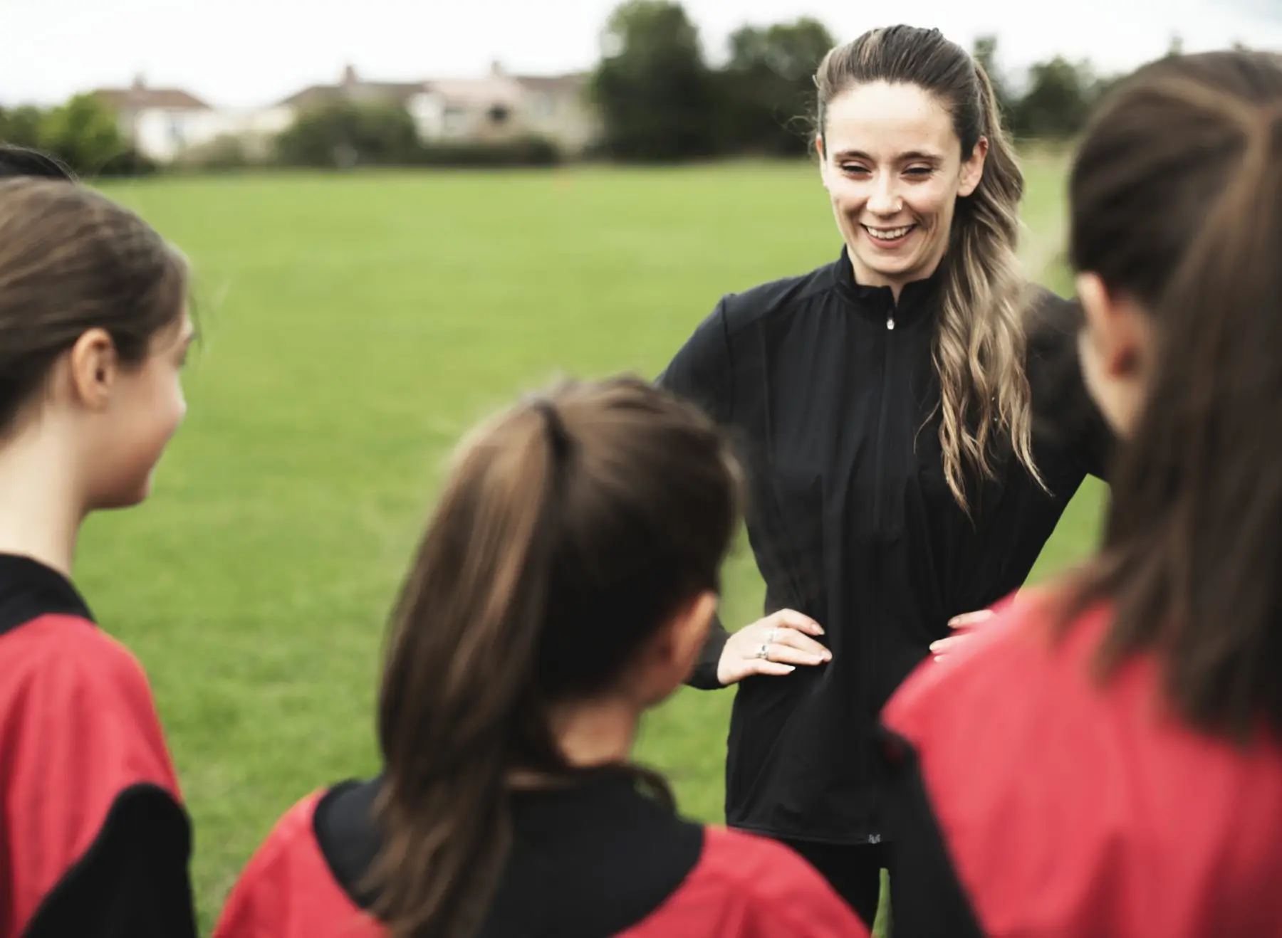 Une entraîneuse souriante, mains sur les hanches, parle à de jeunes joueuses en maillot rouge vues de dos, sur un terrain en herbe