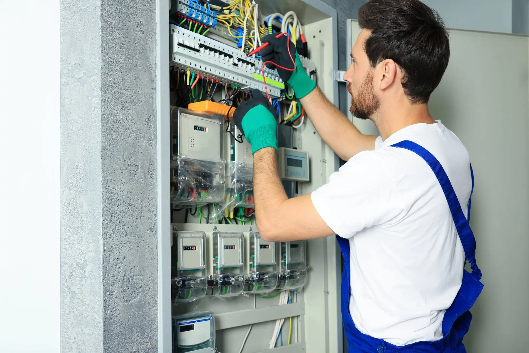 Un électricien en salopette bleue et gants verts mesure au multimètre dans une armoire électrique équipée de plusieurs compteurs