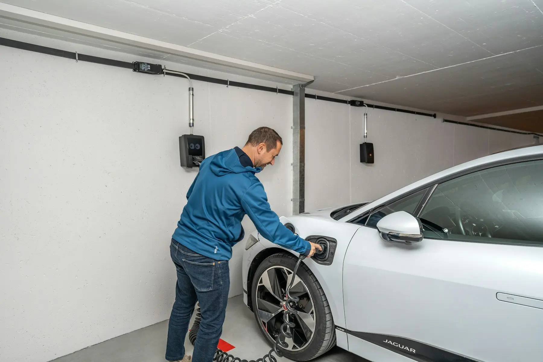 Dans un parking couvert, un homme branche une voiture électrique blanche sur une borne murale ; une seconde borne est fixée plus loin