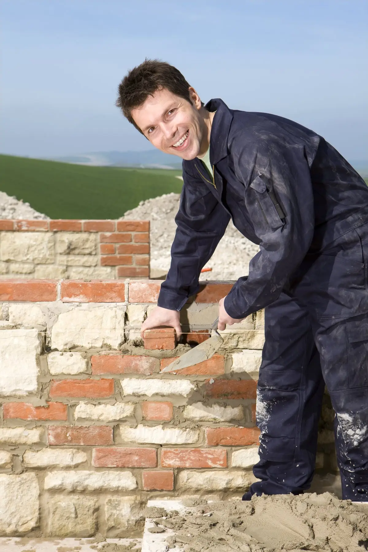 Un maçon en combinaison bleu marine sourit en montant un mur de pierre et de brique, truelle à la main, au milieu des champs