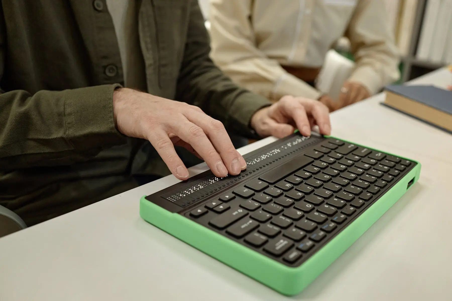 Les mains d’un homme lisent une ligne de braille sur un clavier à plage braille au boîtier vert, posé sur une table
