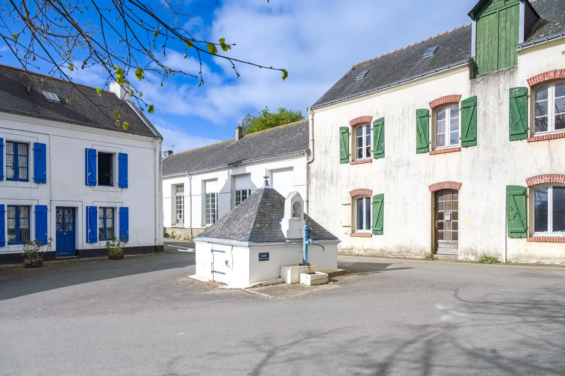 Une petite place de bourg breton : maisons blanches aux volets bleus et verts, toits d’ardoise, et au centre une ancienne fontaine couverte d’un toit d’ardoise