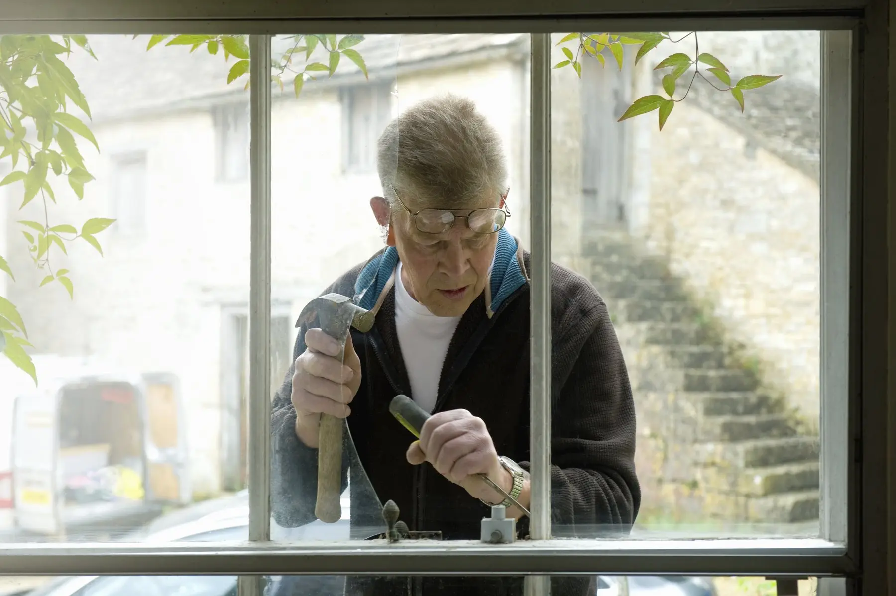 Vu à travers une fenêtre ancienne à petits bois peinte en blanc, un homme âgé répare le châssis, marteau en main ; au fond, une maison et un escalier en pierre