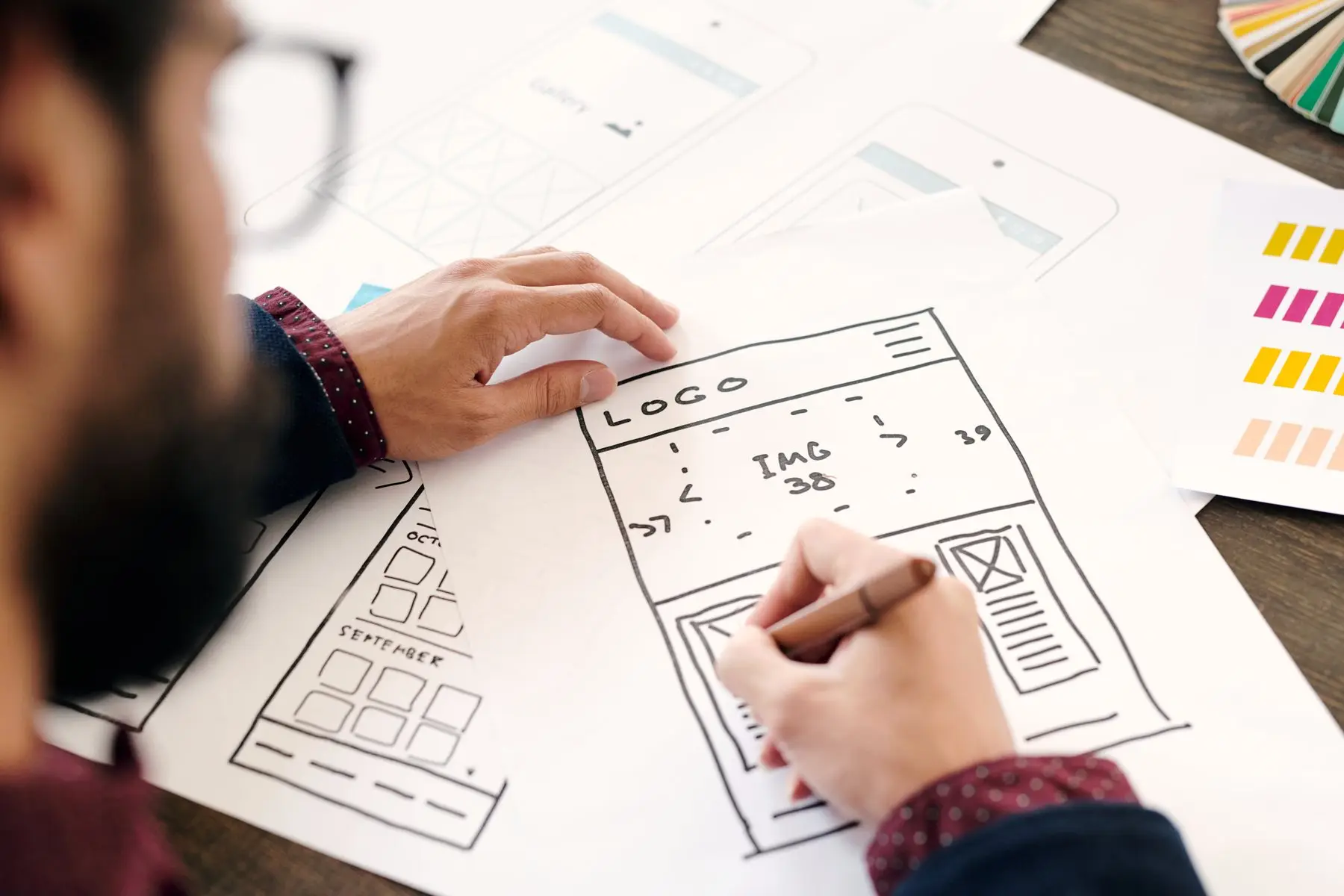Un designer dessine au feutre le plan d’une page web, avec l’emplacement du logo et d’une grande image, à côté d’autres croquis d’écrans et de nuanciers de couleurs