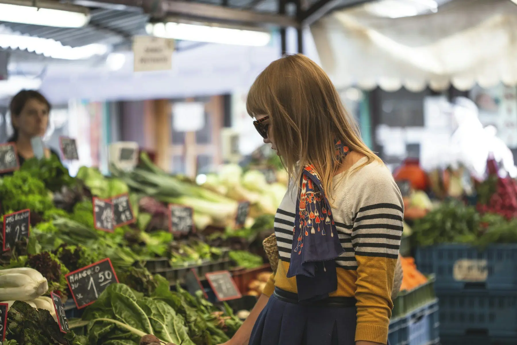 Une cliente devant des étals de légumes et d’herbes aux prix écrits à la craie, sur un marché couvert