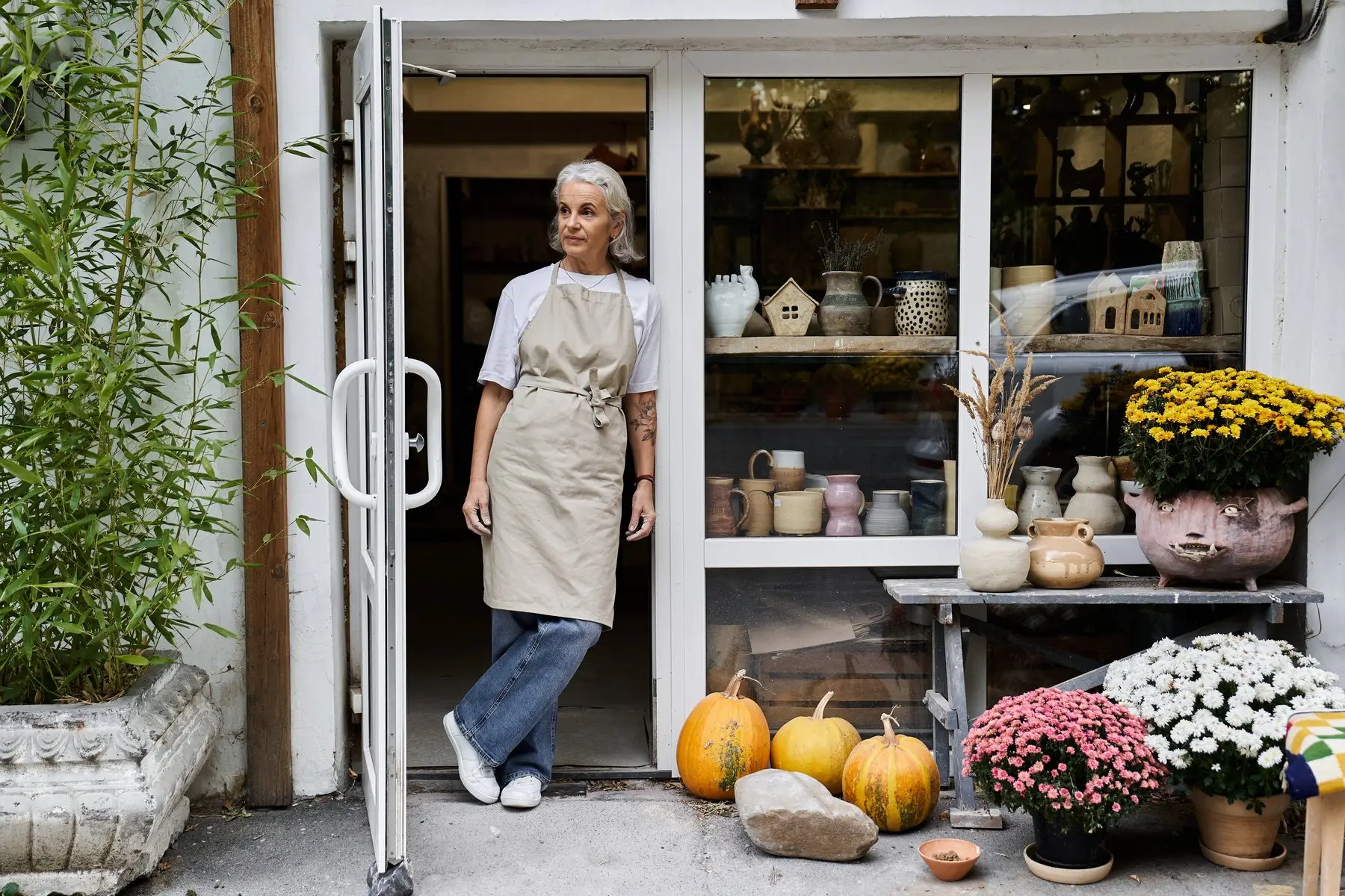 Une céramiste en tablier sur le seuil de sa boutique-atelier, poteries en vitrine et fleurs devant la porte
