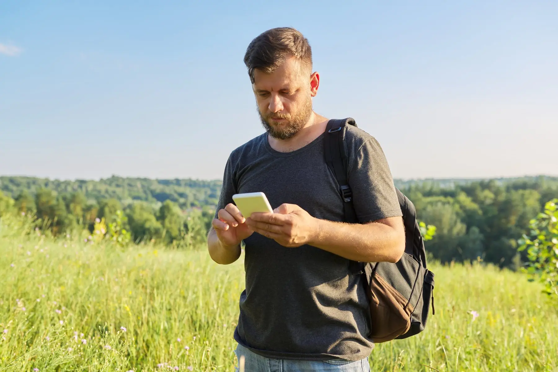 Un randonneur au sac à dos consulte son téléphone au milieu d’une prairie, un jour d’été