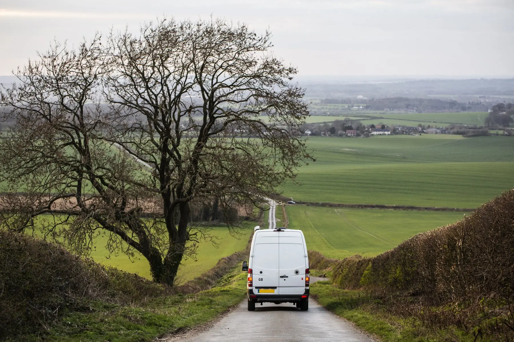 Une camionnette blanche descend une petite route de campagne entre des haies et des champs